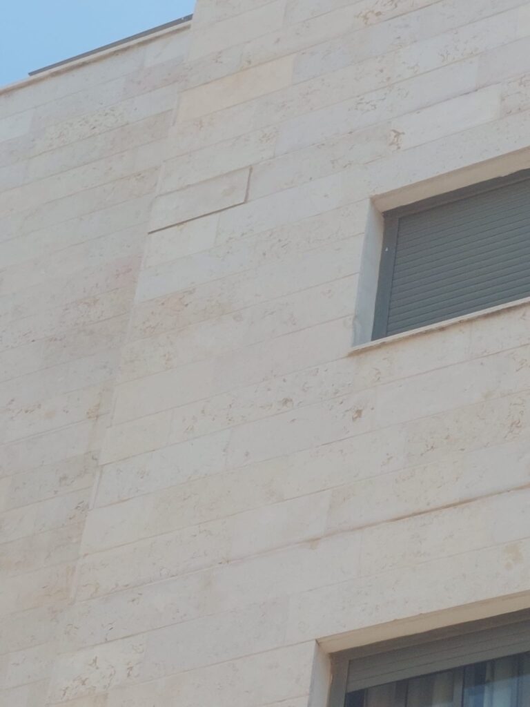 חזית אבן בהירה של בניין מגורים עם לוח אבן רופף ובולט בסמוך לחלון