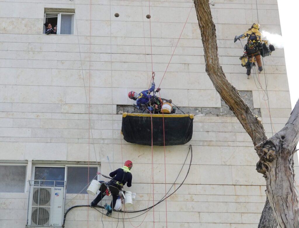 עבודות תיקון חיפוי בחזית בניין עם במה תלויה שחורה ושני גולשים בחבלים מעל יחידות מיזוג