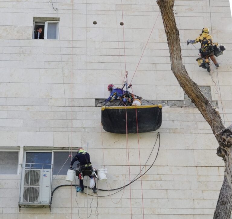 שלושה אנשי צוות על חבלים ובמה תלויה מבצעים שיקום חזית אבן של בניין מגורים