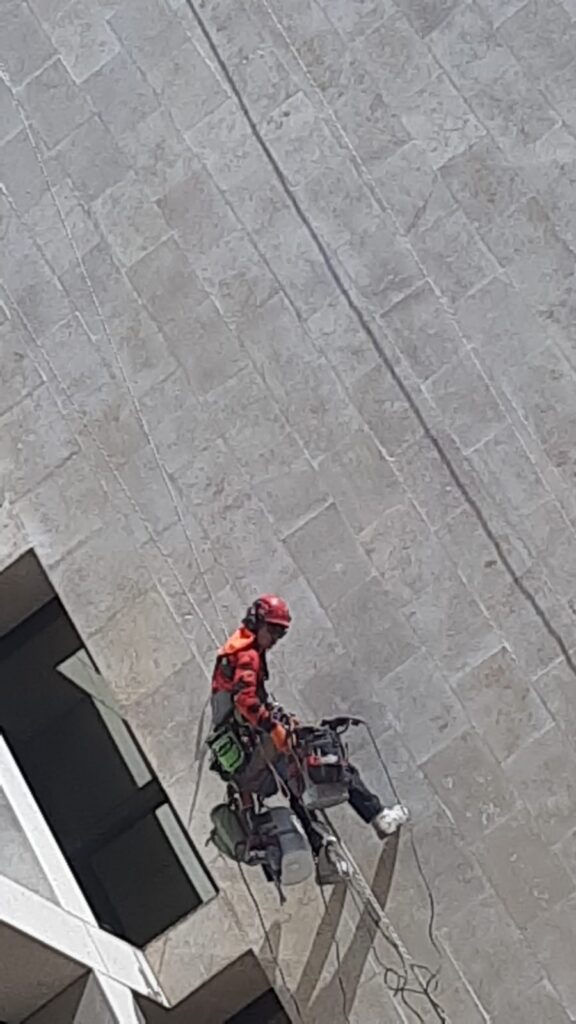 מבט מלמעלה על גולש סנפלינג יושב ברתמה עם ציוד עבודה לאורך חזית מחופה אריחי אבן בהירים