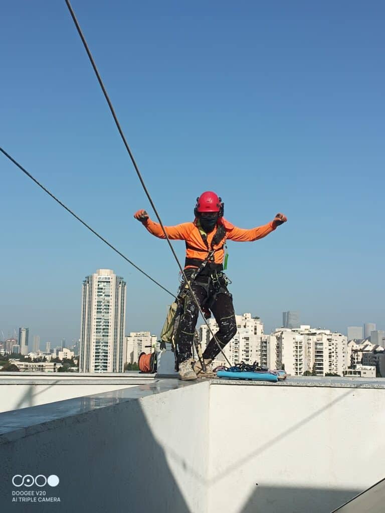 מאמרים עבודות בגובה – בסיס עבודות בגובה עובד גבהים עומד על מעקה גג עם רתמות וחבלים פרוסים על רקע מגדלי מגורים