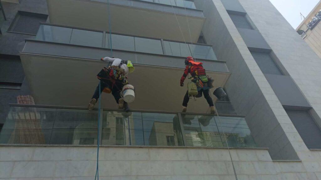 שני גולשי סנפלינג של בסיס תלויים ברתמות מול חזית מגדל מודרני עם מרפסות זכוכית — עבודות איטום ושיקום בגובה ללא פיגומים