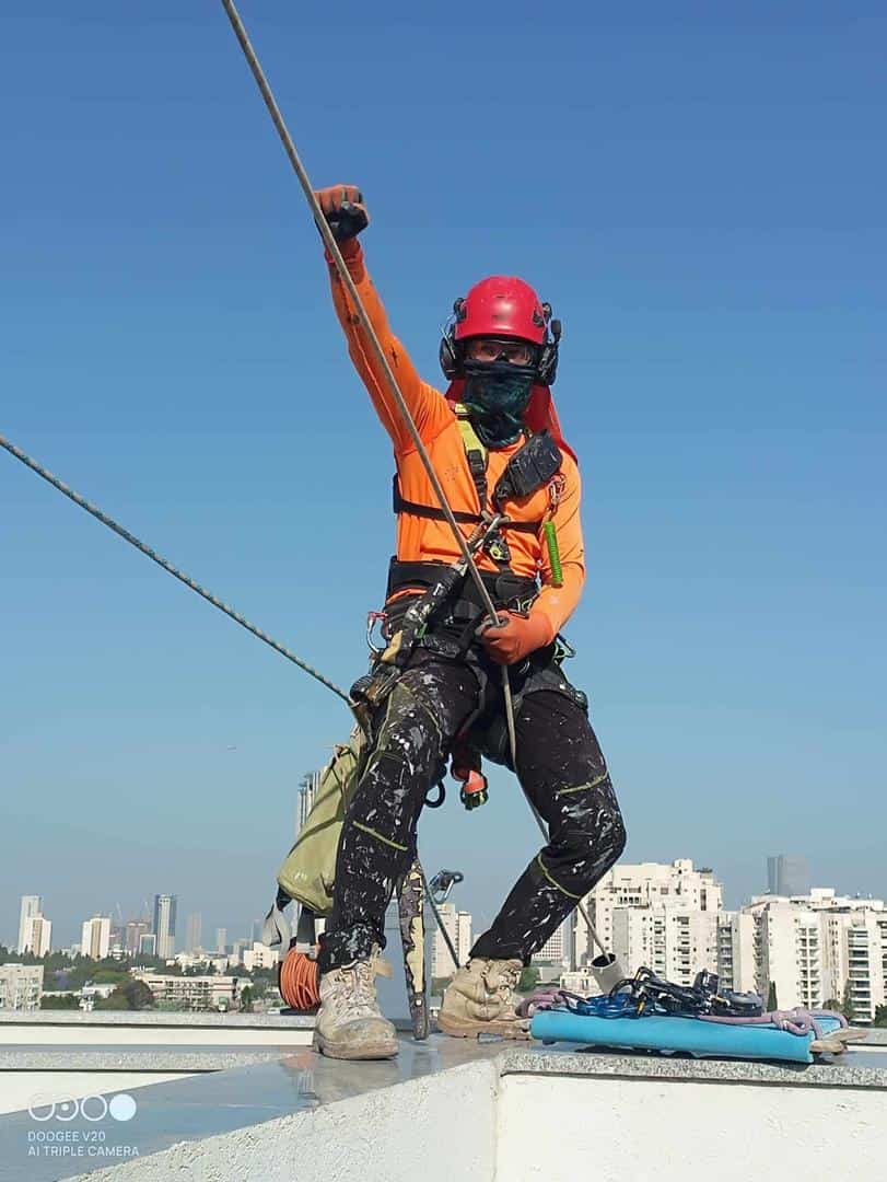 מסנפלן בחולצה כתומה וקסדה אדומה מאובטח בחבלים על מעקה גג מול נוף עירוני